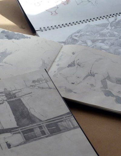 A picture of three sketchbooks on a desk; showing mono pencil drawings of a haberdashery basket, rhinos, and a canal lock.